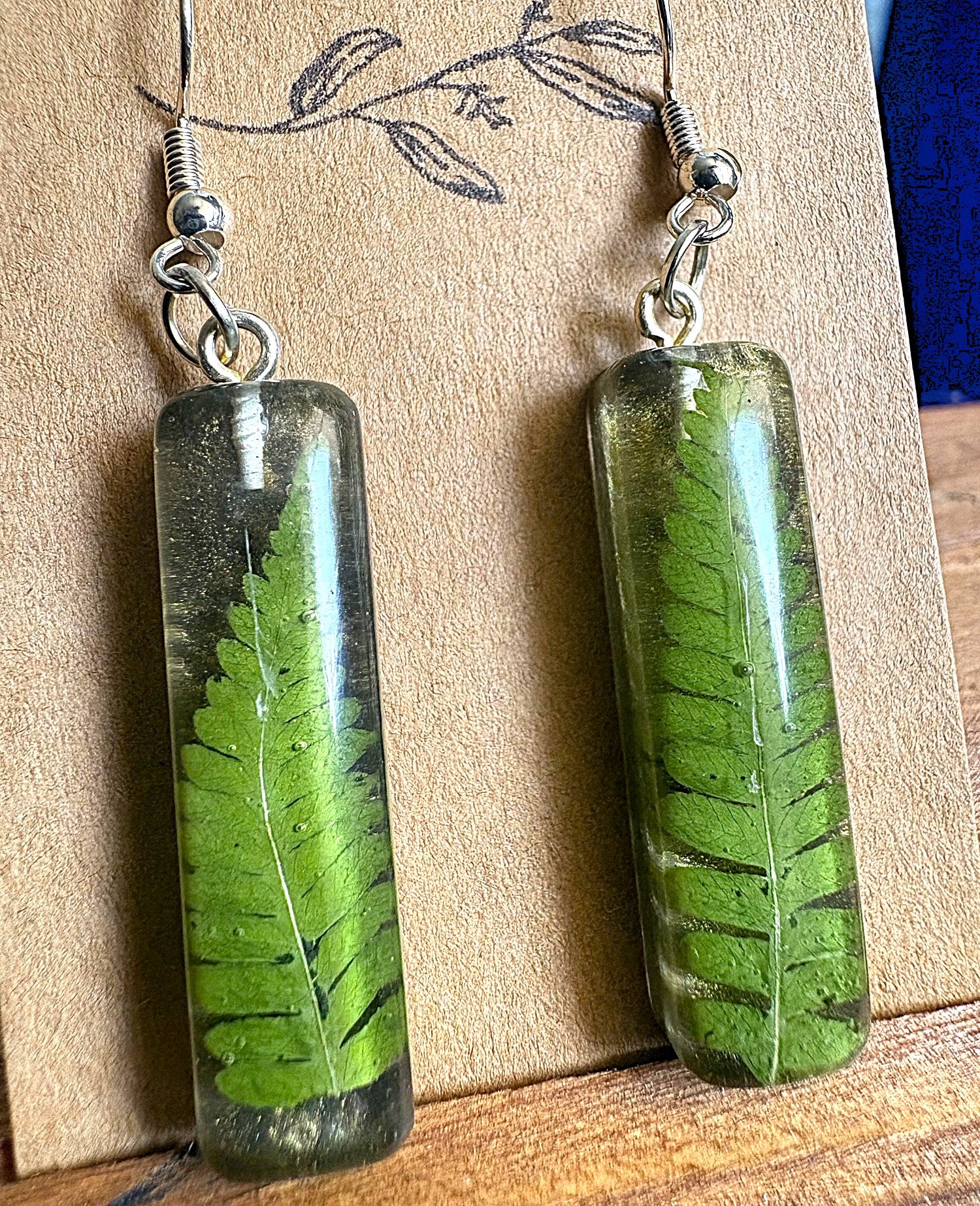 Clear resin bar drop earrings with pressed fern frond displayed on wooden earring card
