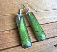 Resin fern earrings lying flat on wood surface showing vivid green fern preserved in clear resin