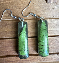 Close-up of resin fern earrings on wood showing detail of pressed fern frond and silver fish hook wires